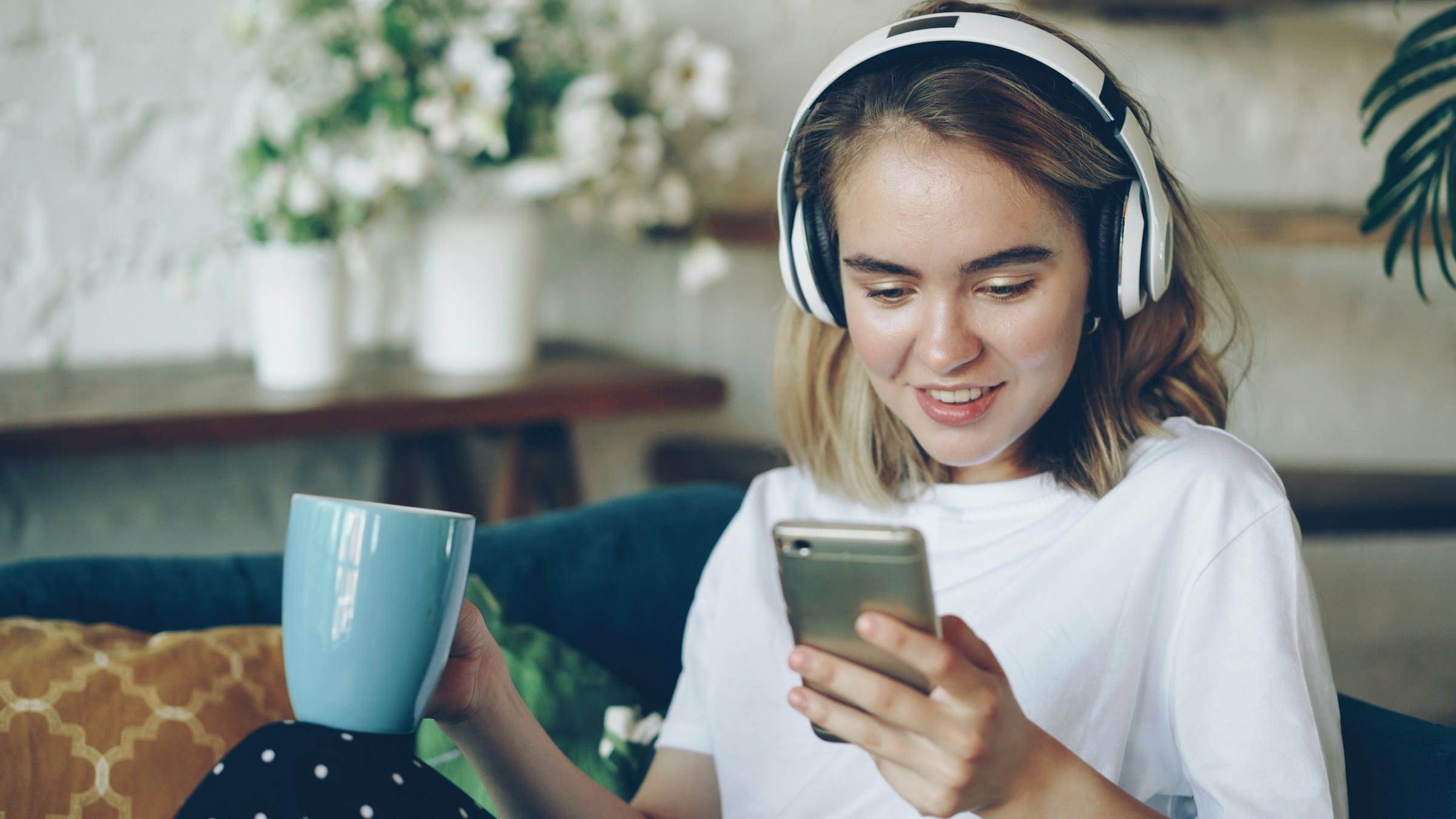 A young woman with headphones and a mug smiling at her phone on the sofa